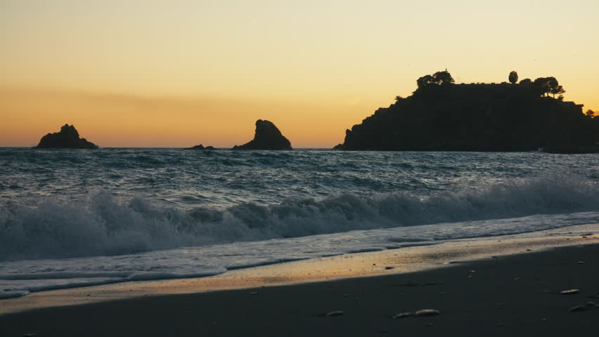 Wide shot of soft evening waves rolling onto shore as the sun sets behind rocky islets and a silhouetted hilltop with trees in southern Spain.