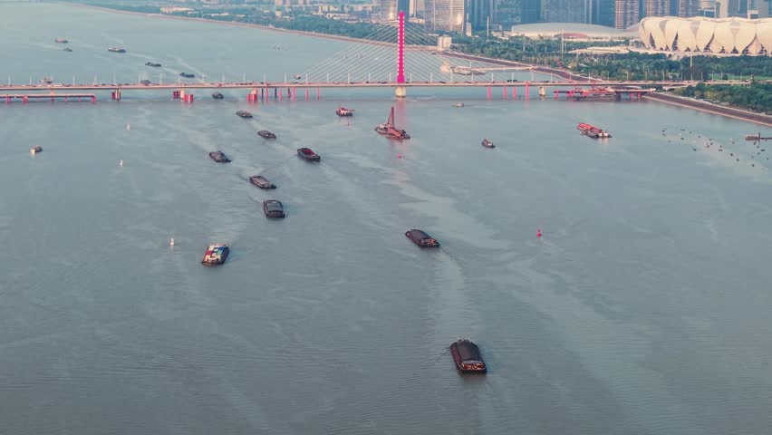 Cargo ships and boats navigate the Qiantang River in Hangzhou China. A red bridge spans the river, with the city skyline visible in the distance