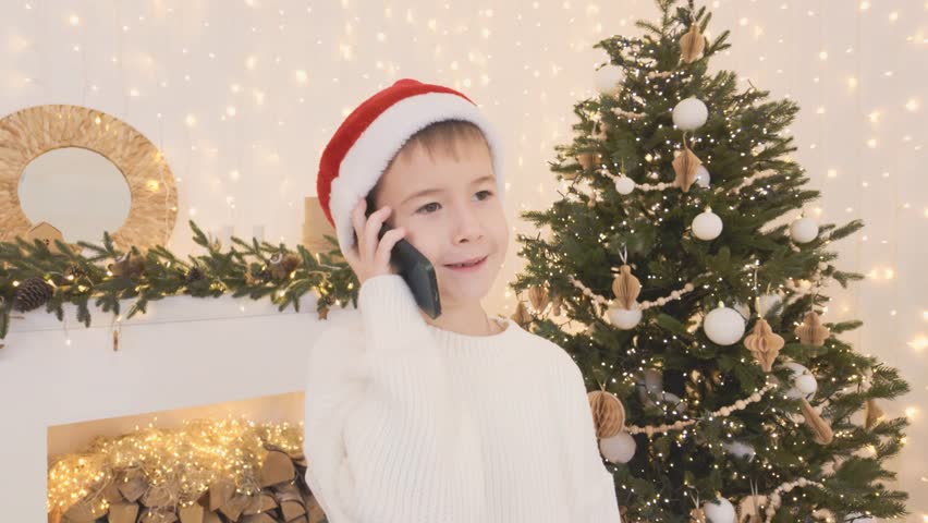 Seven year old boy in white sweater and Santa hat talking on phone call near Christmas tree at home