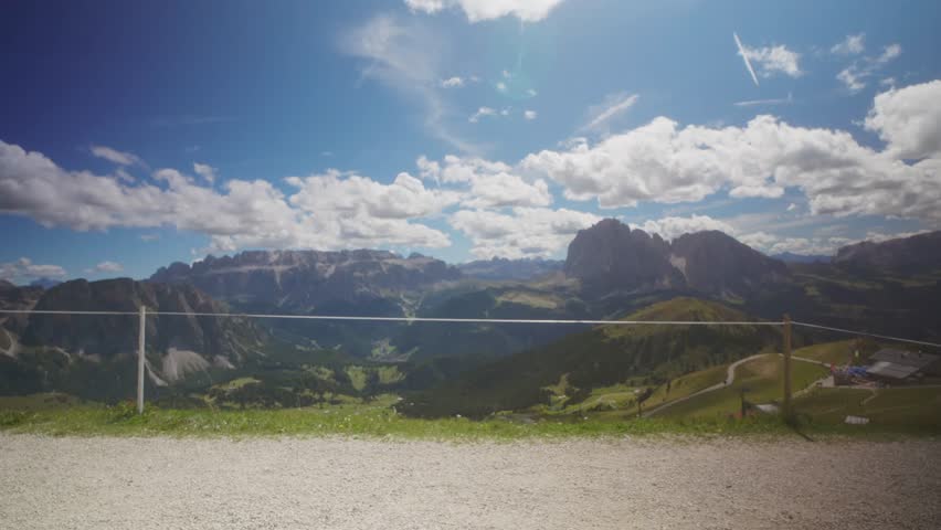 Hiker with backpack walking along a mountain path at Seceda viewpoint in the Dolomites, Italy, with panoramic view of the mountain range under a sunny sky with clouds