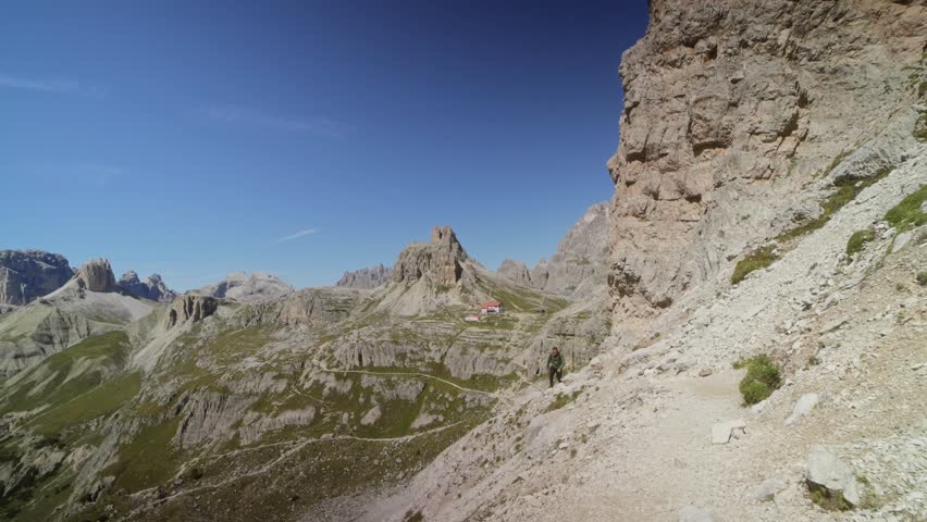 Man walking on hiking path toward Tre Cime di Lavaredo peaks, Dolomites, Italy. Scenic view of alpine mountains and limestone cliffs under clear blue sky