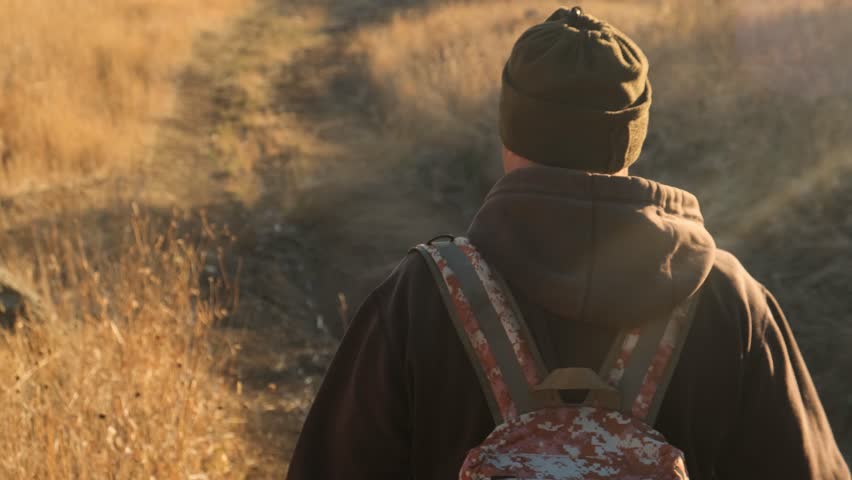 Close-up rear view of a tourist walking through the forest. The traveler carries a camouflage backpack while hiking alone. A walk in the forest in autumn at sunset on a warm day.