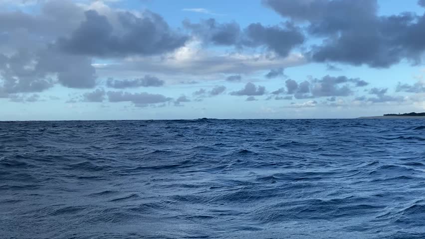 Dynamic landscape video from a boat moving quickly over deep blue choppy water towards a small, lush tropical island under a dramatic cloudy sky