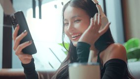 Asian woman smiling happily while listening to music with wireless headphones and holding smartphone, enjoying relaxing moment at cafe, digital lifestyle concept, leisure time, happiness and freedom - Powered by Shutterstock - Get 15% off with code: PIKWIZARD15