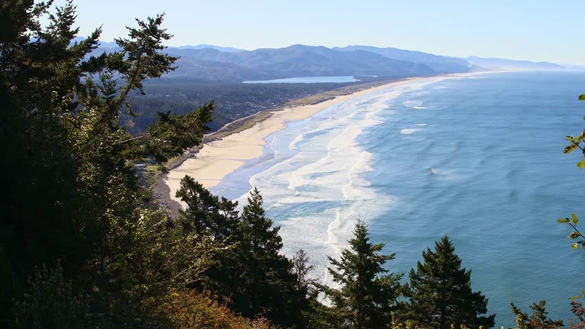An expansive, high-angle aerial view capturing the wide, sandy beach along Tillamook Bay, with the Pacific Ocean and surrounding coastal landscape under a clear, bright, sunny sky in Oregon