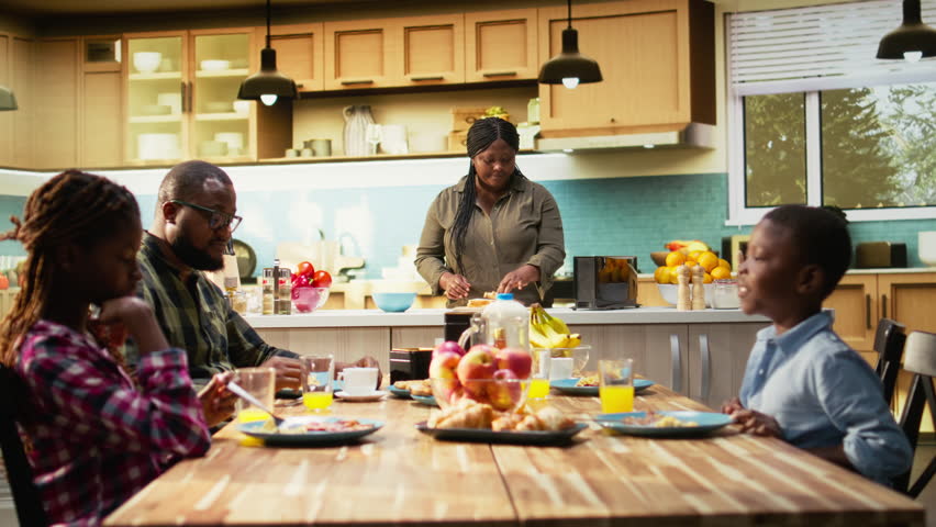 African American mother preparing breakfast in the morning light while the family wait at the table with croissants, fruits and orange juice. Parents and children laugh together, love. Camera A.
