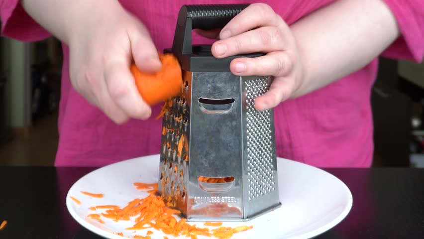 Grating carrots. Hands holding a box grater over a white plate, orange chips, home cooking, close-up.