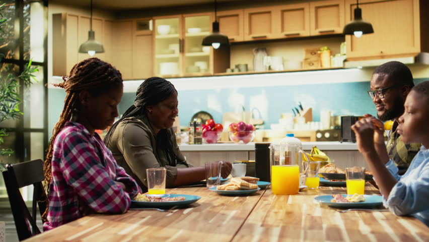 African American family sharing eggs and pastry for breakfast at home, parents serve omelettes, toast and fruit. Cozy kitchen moment captures warmth, relaxation and joyful togetherness. Camera A.