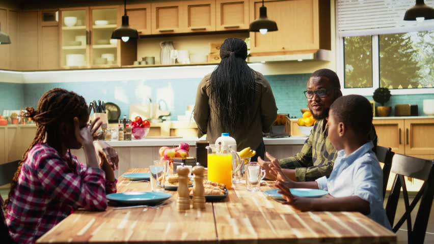 Happy black family enjoying breakfast together in kitchen with delicious food. Mother serves eggs, toast and orange juice while children smile, sharing joy of relaxed weekend morning. Camera A.