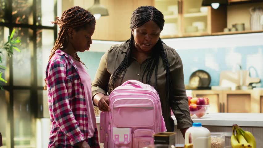 African American mother packing lunch box for her two school kids, preparing their backpack with essentials and snacks for the day. Enjoying rich breakfast with family at home. Camera B.