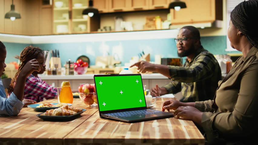 Chroma key next to black family enjoying breakfast at the kitchen table, with plates of omelettes, toast and fruits. Parents and kids laugh together, starting the day with delicious food. Camera A.