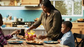African american woman pouring orange juice in glasses at the breakfast table, serving a rich homemade meal with fruits and pastry next to eggs. Cute morning routine for the family. Camera B. - Powered by Shutterstock - Get 15% off with code: PIKWIZARD15