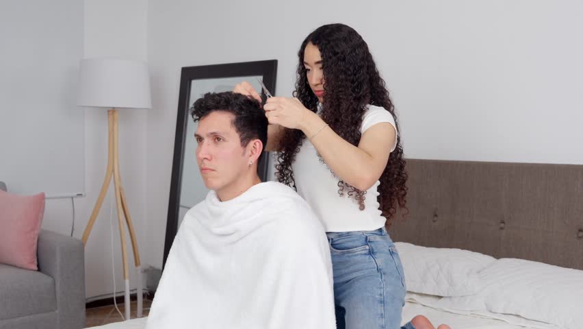 Young girlfriend giving her boyfriend a terrible haircut in their bedroom. Man looks scared and unhappy about his new hairstyle