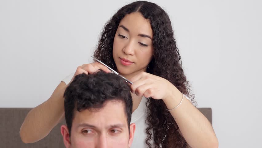 Beautiful latina woman with curly hair giving her boyfriend a haircut at home. Caring relationship and domestic life concept