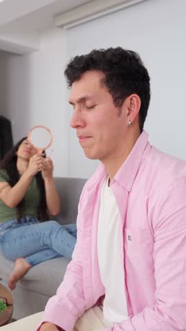 Upset man gesturing and talking heatedly while his indifferent girlfriend sits on the sofa applying lipstick in the background