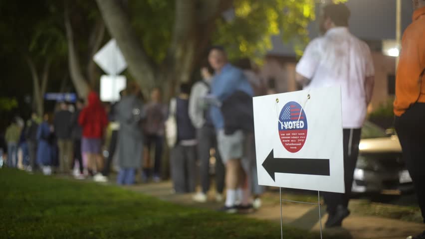 US Elections in Los Angeles, California. "I Voted" sign with voters standing in line on sidewalk in the background as many people are voting late at night.