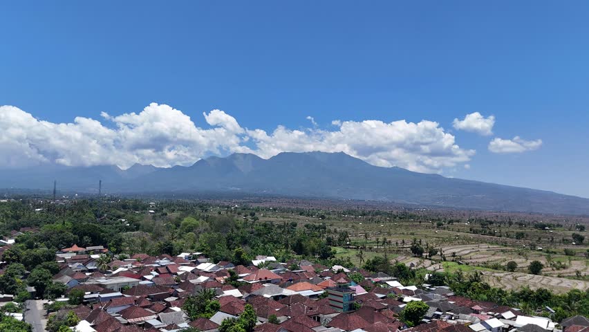 Aerial view of a picturesque mountain village featuring traditional houses, a prominent mosque, lush green trees, and terraced fields under a clear blue sky with fluffy clouds