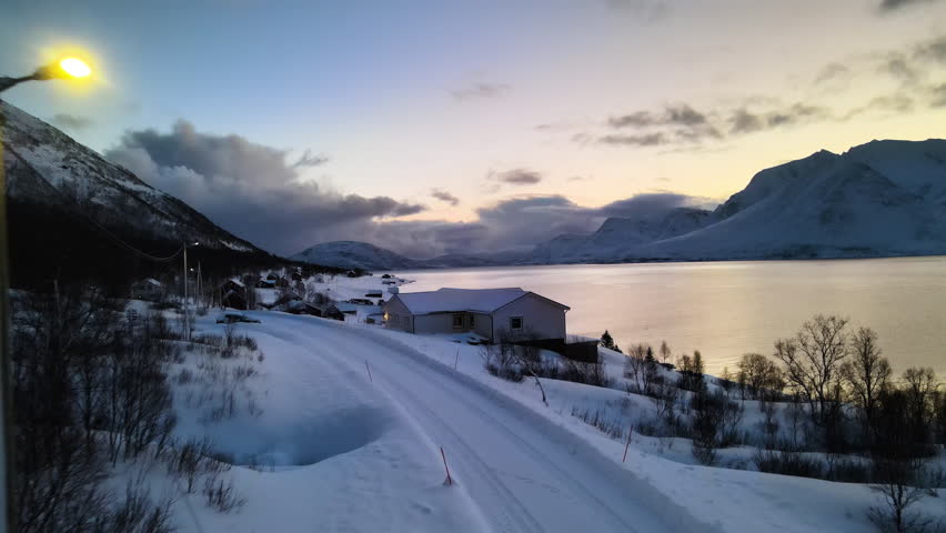 A stunning winter sunset casts warm colors over a snowy landscape, reflecting on the tranquil waters of the fjord near a small village surrounded by mountains.