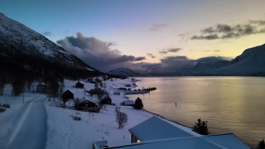 Quiet dawn reveals a stunning snowy landscape along a fjord in Norway. Scenic mountains surround quaint cabins and a peaceful shoreline, creating a serene atmosphere.