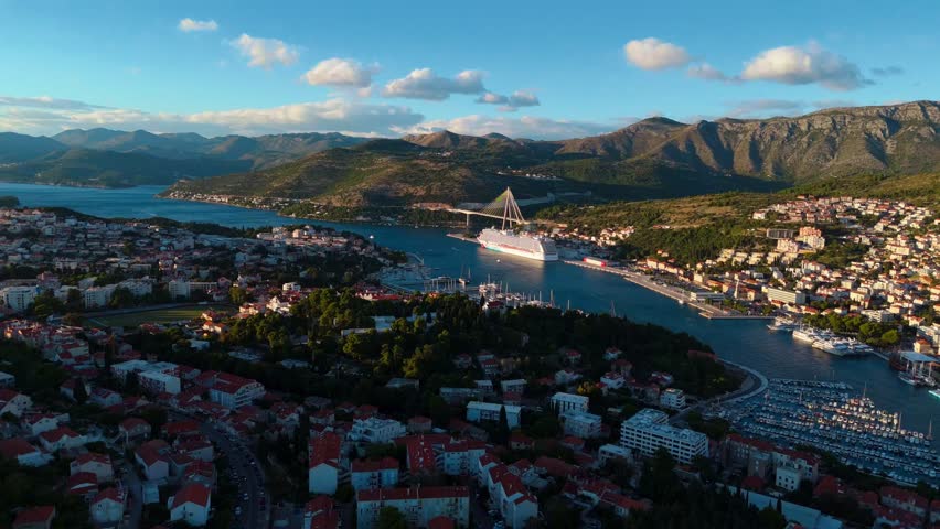 Aerial drone glides toward Dubrovnik’s cruise ship port, revealing docked ships and the dramatic mountain backdrop, all bathed in the warm, golden glow of a sunset over the Adriatic coastline.