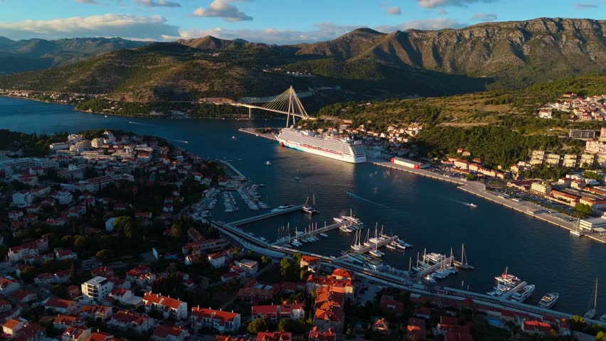 Aerial mid-angle drone glides around Dubrovnik’s cruise ship port, capturing bustling ships, calm waters, and rugged mountains bathed in soft golden sunset light, revealing the city’s coastal charm.