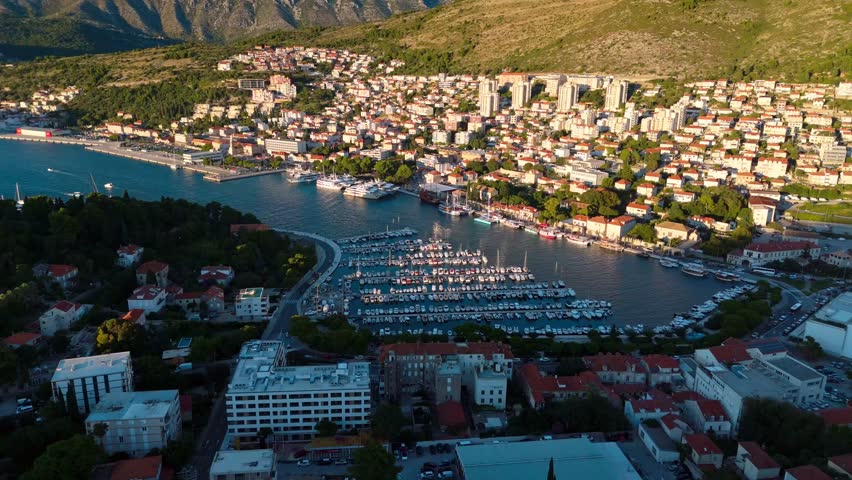 Aerial mid-angle drone soars over Dubrovnik’s cruise ship port and marina, revealing docked ships, waterfront houses, and terracotta rooftops, all glowing warmly under the golden light of sunset.