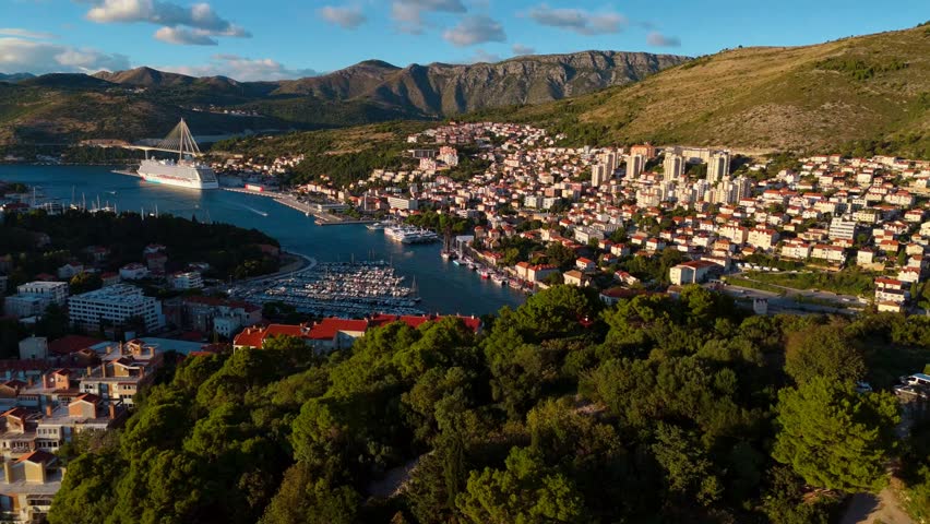 Aerial drone captures a sweeping landscape of Dubrovnik’s coastal town and cruise ship port, with majestic mountains rising in the background, as the warm golden sunset bathes the city and sea