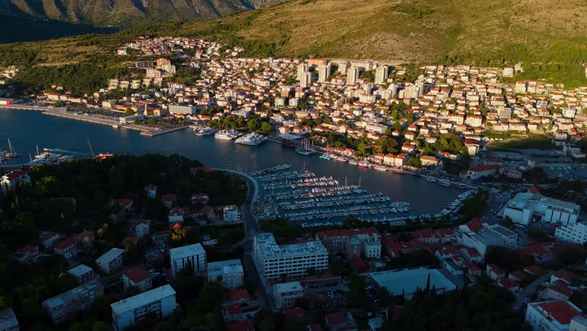 Aerial drone shot flying around Dubrovnik coastal marina port at golden sunset, showcasing rows of docked boats, calm Adriatic waters, and surrounding hills glowing warmly in evening Mediterranean sun