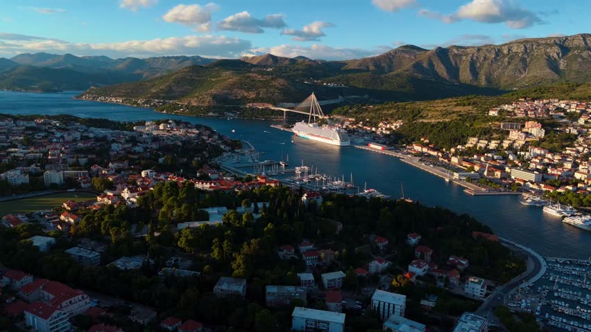 Aerial drone shot circling around Dubrovnik cruise ship port with a mountain backdrop at warm sunset light, showing docked boats, calm Adriatic Sea, and scenic coastal town glowing in evening sun.