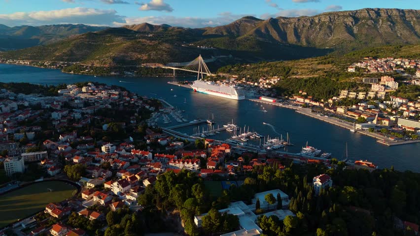 Aerial high angle drone shot flying over Dubrovnik cruise ship port at sunset, showing docked boats and calm Adriatic waters glowing in soft light, surrounded by hills and the coastal city in view