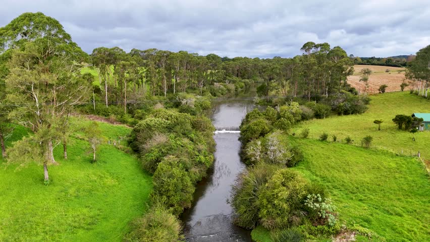 Drone glides above lush farmland, revealing a stream, trees, and concrete weir under cloudy skies