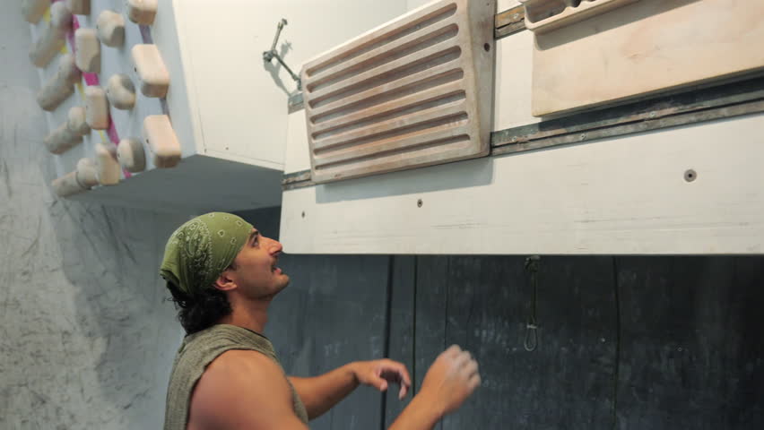 Fixed shot of a man training grip strength on a hangboard, his arms tense with effort as he holds the board and exhales from strain.