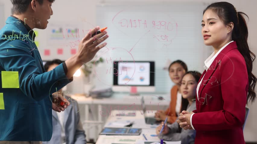 Diverse team of Asian professionals are having a productive meeting in a modern office. Businessman is using a glass whiteboard to present a strategy to team.