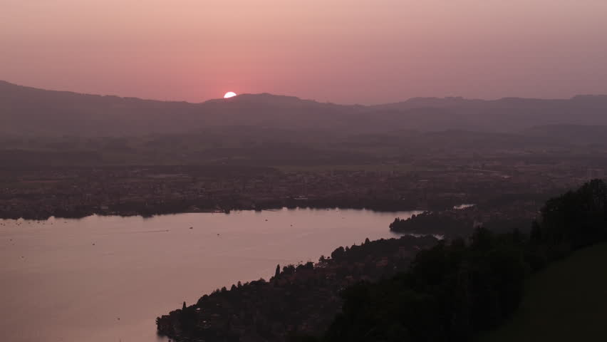 Scenic sunset view over a peaceful mountain lake and valley landscape. Warm sunlight reflects on the water and haze softens the distant hills, evoking calm and natural beauty.