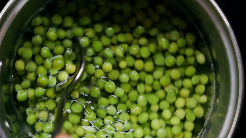Close-up of a spoon stirring green peas in a pot with water, showcasing cooking, fresh ingredients, and meal preparation in a kitchen setting.