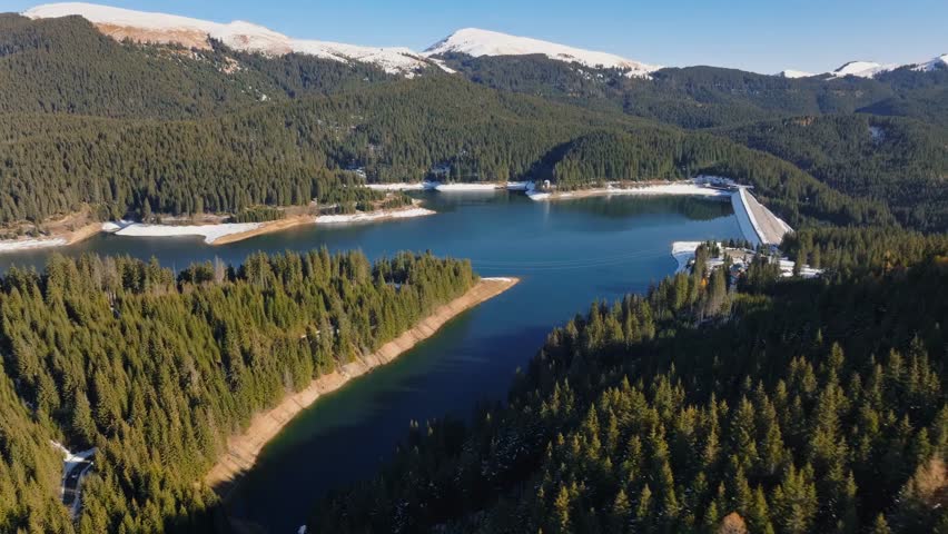 Aerial drone view of Bolboci Lake nestled in the mountains near Brașov, Romania, showcasing serene waters, lush landscapes, and breathtaking natural scenery.