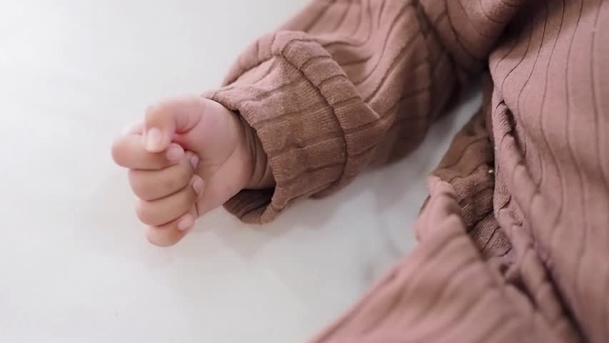 Close up of newborn baby hand is wearing a brown clothes on white rugs