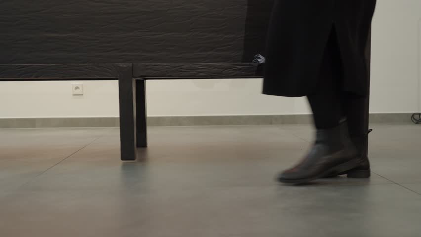 Anonymous figure in dark clothing walks through a minimalist modern art gallery, low angle indoor shot.
