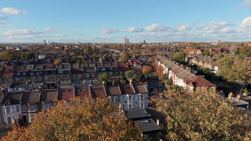 Drone rises slowly above Leyton, East London, showing Victorian houses, autumn trees, and distant tower blocks under blue sky with white clouds. A calm, cinematic establishing shot of the local area.