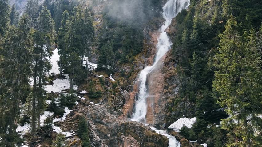 Aerial drone view of Romania’s Carpathian Mountains with a cascading waterfall, lush green forests, and scenic natural landscapes in pristine wilderness.