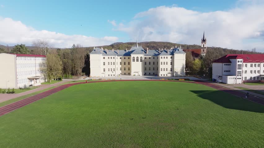 Aerial shot pushing slightly toward the historic Liviu Rebreanu National College building in Bistrita, Romania, positioned behind a bright green athletic field and running track.