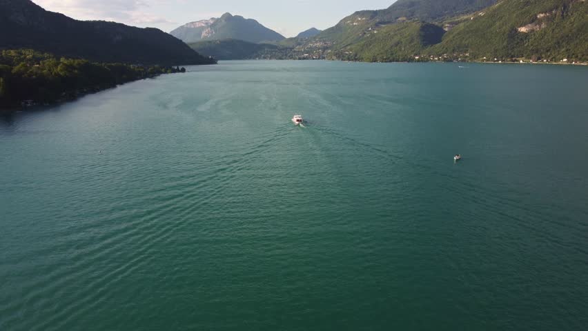 Aerial footage catches up with a ferry travelling along Lake Annecy in France. As the footage catches up with the ferry the drone moves to the right of the ferry as the camera rotates left.