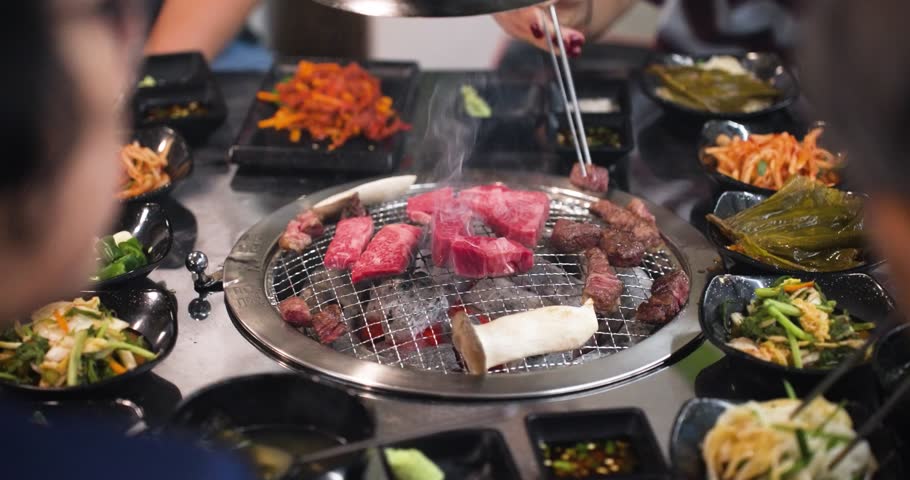 People using chopsticks and enjoying traditional Korean side dishes and noodles as slices of marbled beef sizzle on a round grill in the center of the table