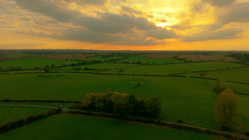 Sunset shining over agricultural fields creating warm golden light. Evening sun illuminating farmland landscape with green meadows. Rural countryside glowing under orange sky