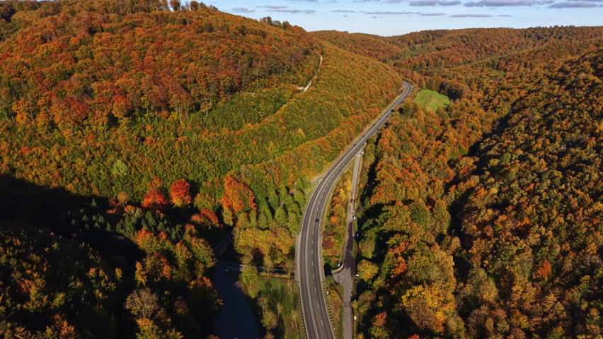 A winding road snakes through a beautiful autumn landscape filled with vibrant orange yellow and red leaves A tranquil river flows alongside creating a picturesque scene.