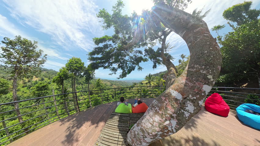 Man and Woman Sitting on a Wooden Bench Under Tree Canopy Overlooking Jungle Landscape with Sunlight, Wide Angle, Relaxing Escape Concept.