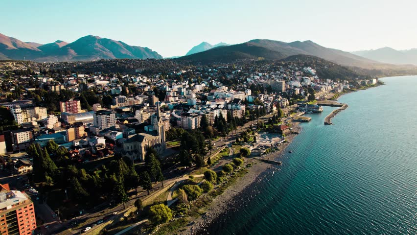 High-angle drone shot capturing the urban core of San Carlos de Bariloche, Argentina, with the historic Cathedral nestled by the vast Nahuel Huapi Lake and the dramatic Patagonian Andes mountains.