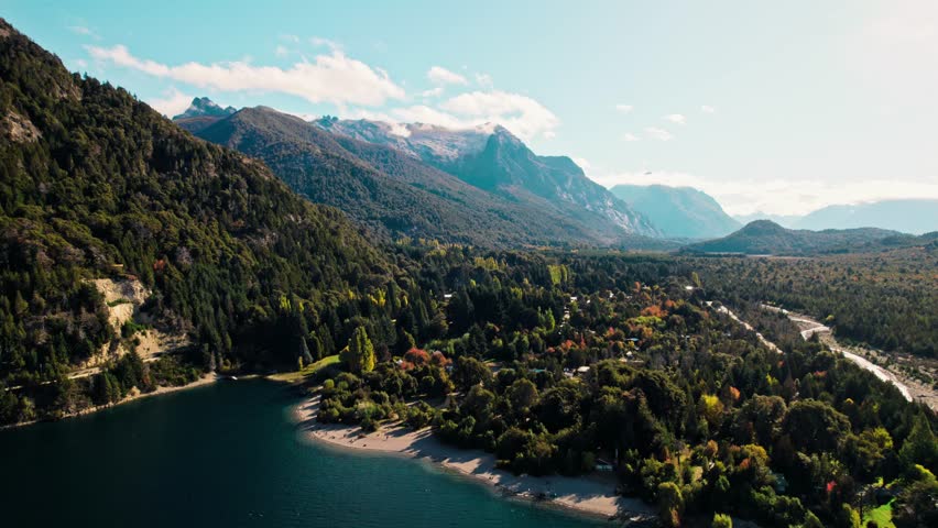 Stunning wide-angle aerial view of the majestic Andes landscape near Bariloche. Captures a deep lake inlet, vast forest slopes, a winding road, and rugged peaks under a bright, clear sky.