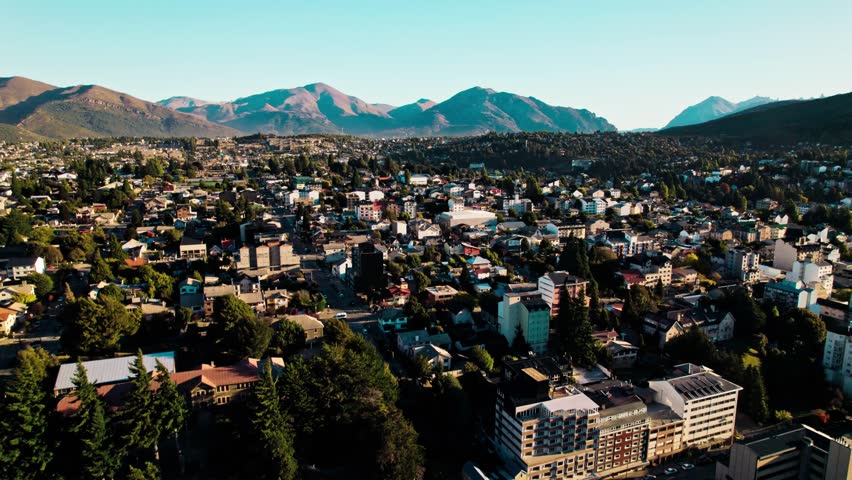Wide-angle drone shot capturing the dense residential areas and urban development of San Carlos de Bariloche, Argentina, contrasting with the vast, rugged slopes of the Patagonian Andes mountains.