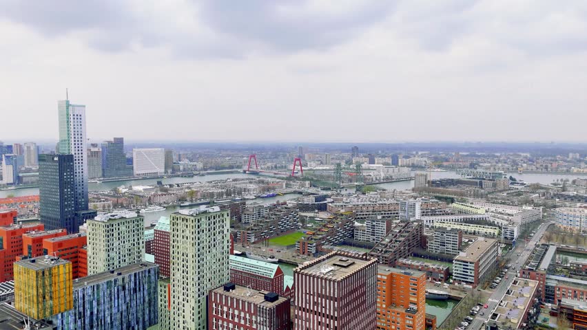 Stunning aerial view of modern Rotterdam skyline featuring Erasmus Bridge, red contemporary buildings, and urban waterfront architecture along Maas River under cloudy daylight sky